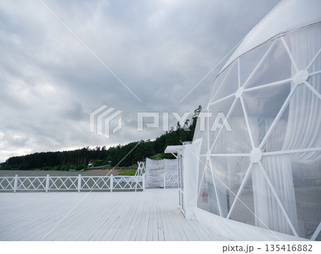 White geodesic dome on wooden deck near lake with forest landscape under cloudy sky. Outdoor glamping tent for event. 135416882