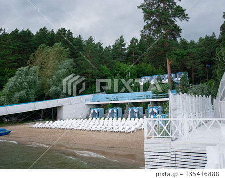 Empty beach with lounge chairs near a green forest and lake shore. Recreation area for summer vacation and travel background. Empty beach with lounge chairs near a green forest and lake shore. Recreation area for summer vacation and travel background. 135416888