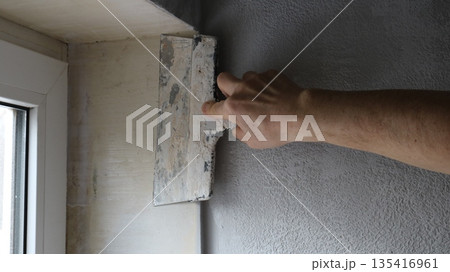 A hand with a wide trowel is leveling the surface of the slope of a small window from inside the room, smoothing the plaster on the window sill with a construction spatula. A hand with a wide trowel is leveling the surface of the slope of a small window from inside the room, smoothing the plaster on the window sill with a construction spatula. 135416961
