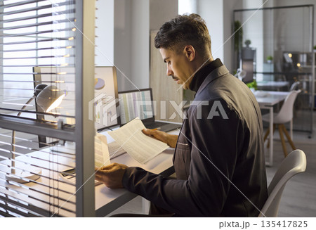 Businessman analyzing printed reports by laptop with graphs on screen in office Businessman analyzing printed reports by laptop with graphs on screen in office 135417825