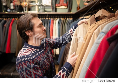 Handsome man consumer choosing clothing in vintage second hand boutique, picking jacket from hanger. Handsome man consumer choosing clothing in vintage second hand boutique, picking jacket from hanger. 135418026