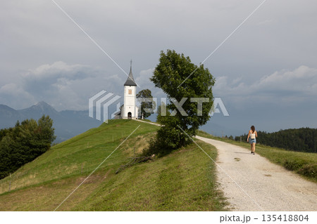Young woman walking towards Church. The Jamnik Church is a charming 15th-century chapel in the Kamnik-Savinja Alps near Kranj, views of the surrounding mountainous landscape. The Church of St. Primoz 135418804