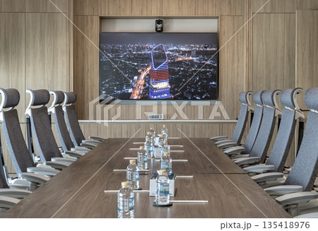 The warm modern boardroom and aesthetic due to the extensive light wood paneling on the walls and the recessed lighting around the screen area. A long, polished wooden table is surrounded by numerous 135418976