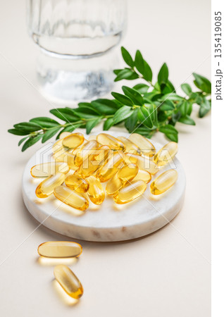Yellow softgel vitamins capsules on a marble stand on a light background with glass of water Yellow softgel vitamins capsules on a marble stand on a light background with glass of water 135419085