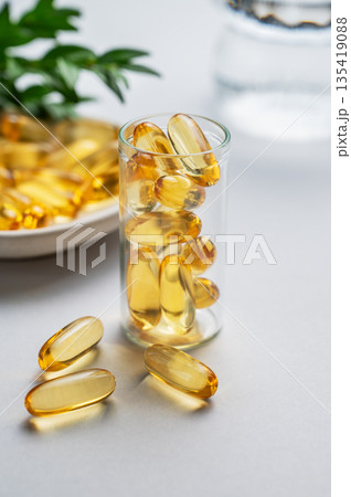 Yellow softgel vitamins capsules scattered in a bottle on a light isolated background with branch. Yellow softgel vitamins capsules scattered in a bottle on a light isolated background with branch. 135419088