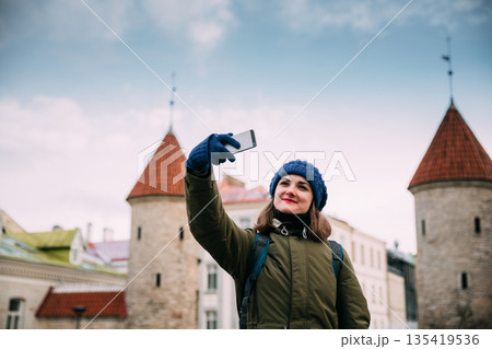 Tallinn, Estonia. Young Caucasian Woman Backpacker Tourist Enjoying Life, Smiling And Using Smartphone Near Landmark Viru Gate. Vacation In Old Town. Girl Surfing Net Internet In Phone 135419536