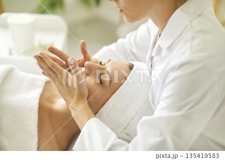 Close up masseur hands making face relaxing massage to a young girl in beauty salon. Close up masseur hands making face relaxing massage to a young girl in beauty salon. 135419583