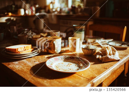 The table shows two empty cups and a plate with crumbs. There is also a stack of bread on a plate in a kitchen with morning light shining through the window 135420566