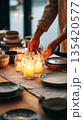 A person is placing simple candles inside jars on a dining table. The setup takes place in a casual home kitchen during the evening and creates a cozy atmosphere 135420577