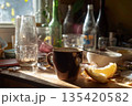 A breakfast table is set with a cup, scattered food scraps, and empty bottles and glasses. The sunlight shines through the window, creating warm shadows on the table 135420582