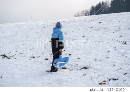 Active little boy in warm winter clothes holding a blue sled while walking uphill on a snowy slope, enjoying outdoor winter fun and play Active little boy in warm winter clothes holding a blue sled while walking uphill on a snowy slope, enjoying outdoor winter fun and play 135421094