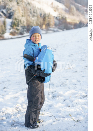 Happy child dressed in warm winter clothes holding a blue sled on a snowy hillside, enjoying outdoor winter activities and family leisure time. 135421095