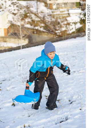 Active little boy in warm winter clothes holding a blue sled while walking uphill on a snowy slope, enjoying outdoor winter fun and play. 135421097