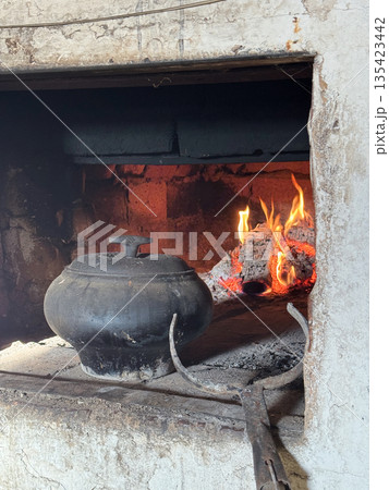 Cast iron covered with a lid on the hearth of a Russian stove. A stove hook is visible nearby. In the background, fire burns in the stove firebox. Cooking food in a vintage stove. Cast iron covered with a lid on the hearth of a Russian stove. A stove hook is visible nearby. In the background, fire burns in the stove firebox. Cooking food in a vintage stove. 135423442