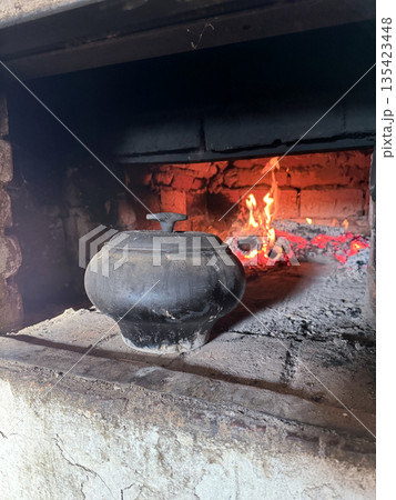 Cast iron covered with a lid. Stands on the hearth of a Russian stove. In the background, fire burns in the stove firebox. Cooking food in a vintage stove. Cast iron covered with a lid. Stands on the hearth of a Russian stove. In the background, fire burns in the stove firebox. Cooking food in a vintage stove. 135423448