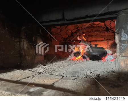 Cast iron covered with a lid. Stands in the stove firebox. Among coals and burning firewood. Cooking food in a Russian stove. Cast iron covered with a lid. Stands in the stove firebox. Among coals and burning firewood. Cooking food in a Russian stove. 135423449