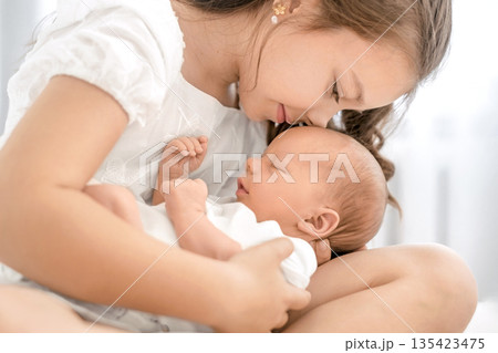 The older sister kisses the younger newborn brother, holding him gently and carefully in arms. The older sister kisses the younger newborn brother, holding him gently and carefully in arms. 135423475
