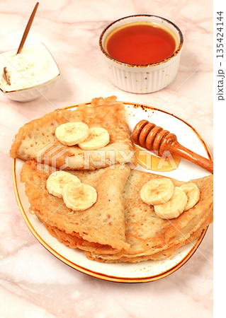 Delicious Russian pancakes with soft cheese, bananas, and honey on a marble table. A healthy breakfast made with fresh ingredients, inspired by Maslenitsa and traditional Russian cuisine 135424144