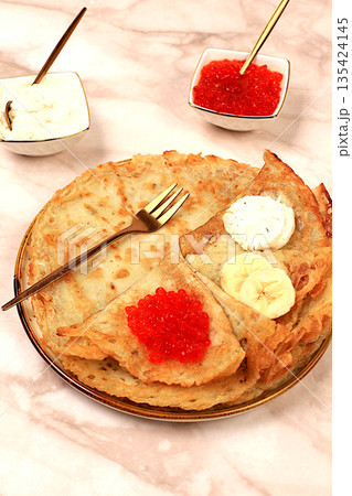 Delicious Russian pancakes with soft cheese, bananas, and red caviar on a marble table. Healthy breakfast with fresh ingredients, Maslenitsa concept and traditional Russian cuisine, selective focus Delicious Russian pancakes with soft cheese, bananas, and red caviar on a marble table. Healthy breakfast with fresh ingredients, Maslenitsa concept and traditional Russian cuisine, selective focus 135424145