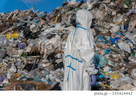 A worker in protective clothing inspects a large area of trash at a landfill, surrounded by plastic and waste materials A worker in protective clothing inspects a large area of trash at a landfill, surrounded by plastic and waste materials 135424442