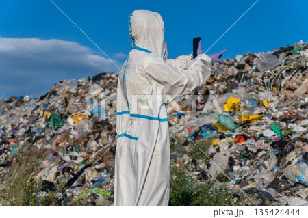 A person wearing a protective suit and gloves points at a large mound of garbage in a landfill site while clouds drift in the blue sky 135424444