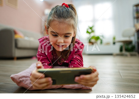 Young girl immersed in digital adventure at home, exploring a colorful game on her phone while relaxing on a cozy rug during a warm afternoon 135424652