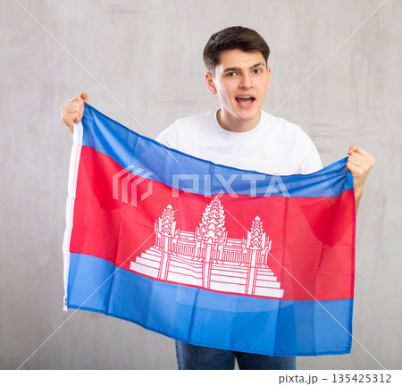 Energized,active male spectator holds national flag of Cambodia in hands and chants popular slogan. 135425312