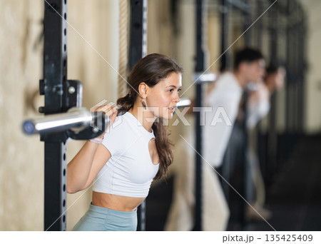 Girl in activewear exercising with barbell during group workout in gym center Girl in activewear exercising with barbell during group workout in gym center 135425409