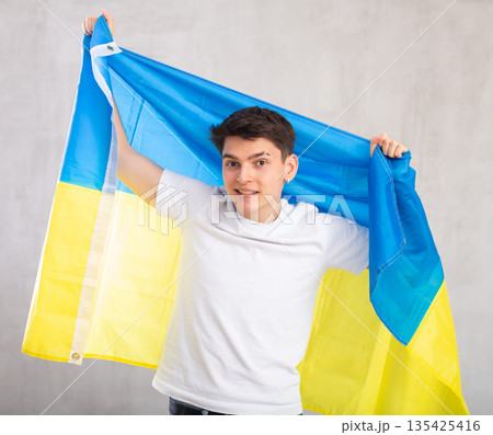 Reliant positive young man holds big national flag of Ukraine with confident smile showing teeth. Reliant positive young man holds big national flag of Ukraine with confident smile showing teeth. 135425416