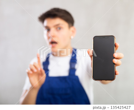 Unfocused man in jumpsuit shows close up empty mobile phone screen and points at device with finger 135425427