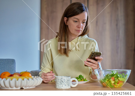 Woman sitting at table using smartphone while eating fresh salad 135425661