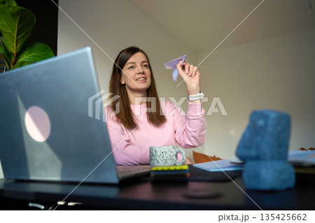Woman sitting at desk holding paper airplane and looking away from laptop Woman sitting at desk holding paper airplane and looking away from laptop 135425662