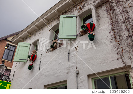 Three mint shuttered windows display teddy bears peeking and floating in balloon baskets, adding playful charm to a festive white facade with string lights and vines. Three mint shuttered windows display teddy bears peeking and floating in balloon baskets, adding playful charm to a festive white facade with string lights and vines. 135426206