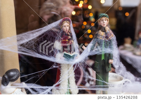 Two Victorian style caroler figurines covered in cobwebs stand near mugs and festive lights, evoking eerie nostalgia. 135426208