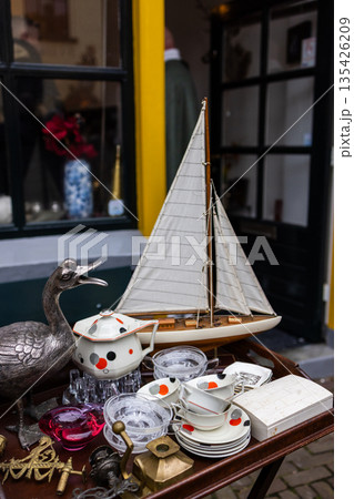 Outdoor table displays model sailboat with white sails, ceramic dishes with spiral motifs, glassware, and metal bird sculpture near shop entrance. Outdoor table displays model sailboat with white sails, ceramic dishes with spiral motifs, glassware, and metal bird sculpture near shop entrance. 135426209