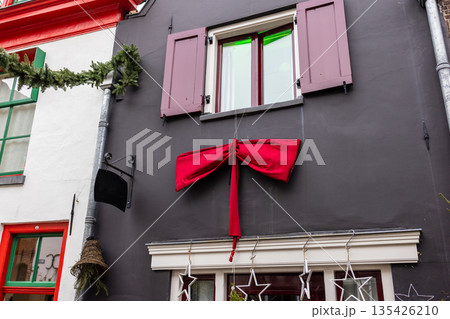 Dark gray facade with maroon shutters and red bow, festive stars and garlands adorn windows and roofline in holiday street scene. Dark gray facade with maroon shutters and red bow, festive stars and garlands adorn windows and roofline in holiday street scene. 135426210