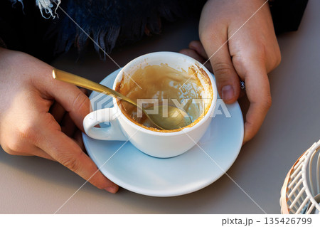Hands holding an almost empty coffee cup on a saucer with a spoon, with visible latte residue in warm sunlight at a cafe table. 135426799