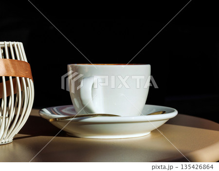 Sunlit cafe table with white coffee cup on a saucer with a spoon against a black background with large black copy space Sunlit cafe table with white coffee cup on a saucer with a spoon against a black background with large black copy space 135426864