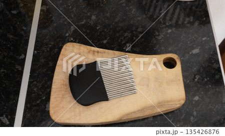 Overhead view of a stainless steel crinkle cutter on a wooden chopping board. Kitchen utensil with wavy blade for slicing vegetables. Black granite countertop background Overhead view of a stainless steel crinkle cutter on a wooden chopping board. Kitchen utensil with wavy blade for slicing vegetables. Black granite countertop background 135426876