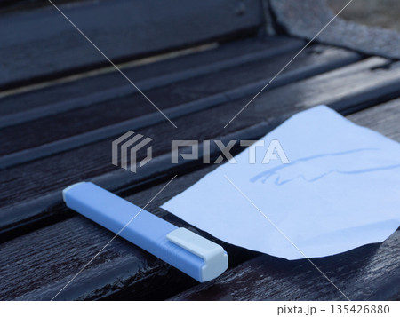 Blue marker pen and white paper with a scribble on a wooden park bench. Close-up of stationery and a doodle on a textured surface 135426880