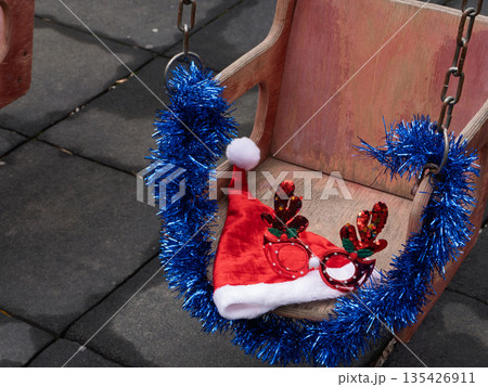 Christmas decorations on a wooden swing in a playground. Red Santa hat and glittery reindeer glasses with blue tinsel. Festive holiday season outdoors Christmas decorations on a wooden swing in a playground. Red Santa hat and glittery reindeer glasses with blue tinsel. Festive holiday season outdoors 135426911