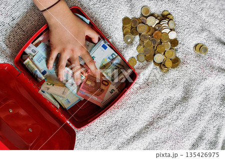 Overhead view of a young woman hand touching euro banknotes inside an open red cash box and a pile of euro coins on a bed blanket Overhead view of a young woman hand touching euro banknotes inside an open red cash box and a pile of euro coins on a bed blanket 135426975