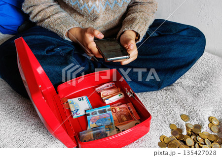 Young woman on bed using smartphone and counting euro cash from open cash box Young woman on bed using smartphone and counting euro cash from open cash box 135427028