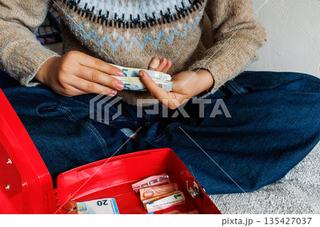 Young woman sitting on bed counting euro banknotes beside open red cash box 135427037