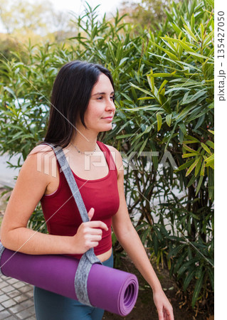 Young woman carrying yoga mat outdoors, walking for wellness Young woman carrying yoga mat outdoors, walking for wellness 135427050