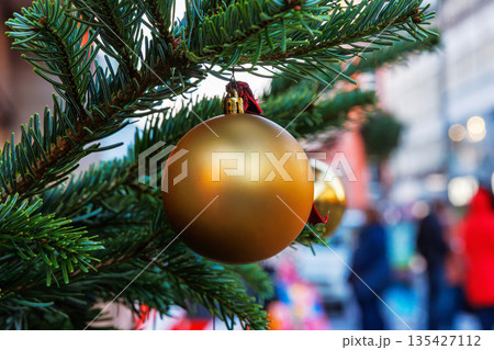 A gold christmas bauble hanging from a fir branch with a blurred street scene and people in the background. 135427112