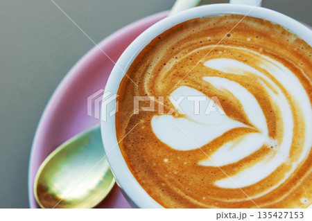 A closeup top view of a cappuccino topped with heart and flower shaped latte art in a white cup on a pink saucer with  a small spoon beside. Soft focus 135427153