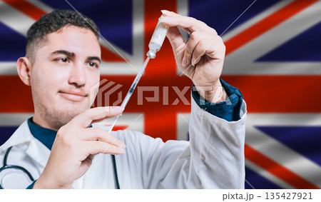 Doctor holding syringe on United Kingdom flag background. Medical professional holding syringe with British flag in the background Doctor holding syringe on United Kingdom flag background. Medical professional holding syringe with British flag in the background 135427921