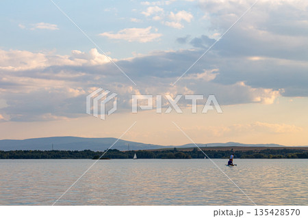 Paddleboarder enjoys a calm evening on the water with scenic mountains in the background during sunset 135428570