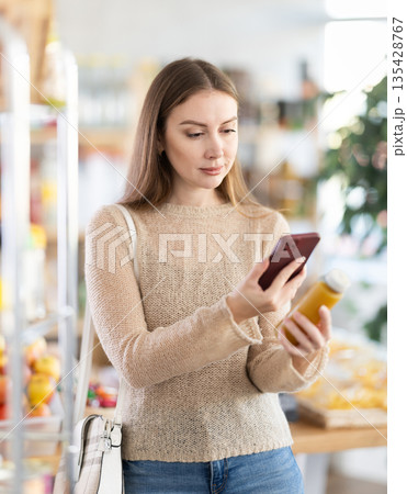Young attentive girl buyer in casual clothes in supermarket scan QR code bottle of yellow smoothie Young attentive girl buyer in casual clothes in supermarket scan QR code bottle of yellow smoothie 135428767